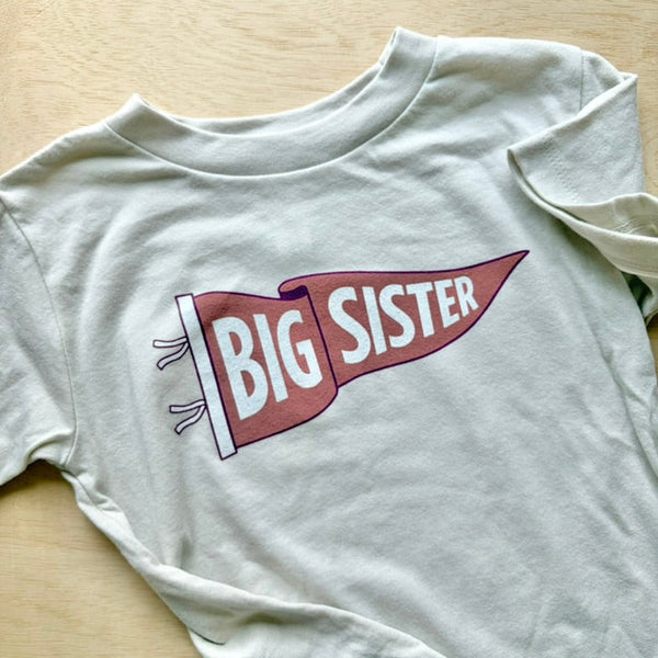 A light gray long-sleeve t-shirt laid flat on a light wood surface, featuring a graphic of a coral and white pennant flag with the words 'BIG SISTER' in white.