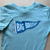 A light blue short-sleeve t-shirt laid flat on a light wood surface, featuring a blue and white graphic print on the chest that reads 'BIG BROTHER' inside a pennant design.